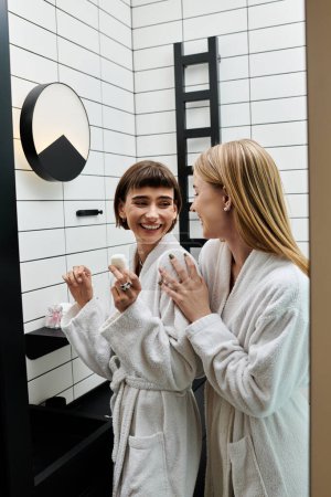 Two young women in bath robes standing in front of a bathroom mirror, gazing at each other with affection.
