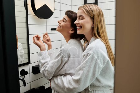 A couple of women in bath robes stand together, sharing a tender moment in front of a bathroom mirror.