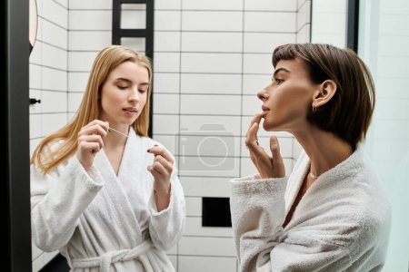 A young woman in a bathrobe flossing her teeth while looking at her partner in a hotel bathroom.