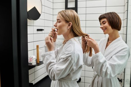 A young lesbian couple in bath robes, one flossing teeth, other braiding hair in a hotel bathroom.