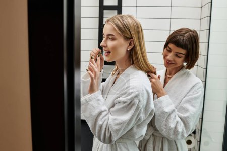 Zwei junge Frauen, ein lesbisches Paar, stehen in einem Hotelbadezimmer vor einem Spiegel