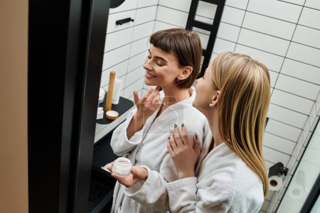 Zwei Frauen in Bademänteln stehen in einem Hotelbadezimmer vor einem Spiegel und teilen einen zärtlichen Moment der Besinnung und Intimität.