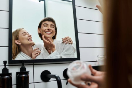 Foto de A young women in a bath robes stands in front of a mirror, applying cream in a hotel bathroom. - Imagen libre de derechos