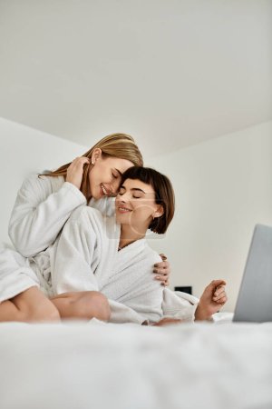 Photo for A young lesbian couple in bath robes relaxes on top of a cozy bed in a hotel room. - Royalty Free Image