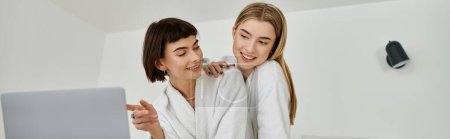 Photo for Young lesbian couple in bath robes looking intently at a laptop screen inside a hotel bathroom. - Royalty Free Image