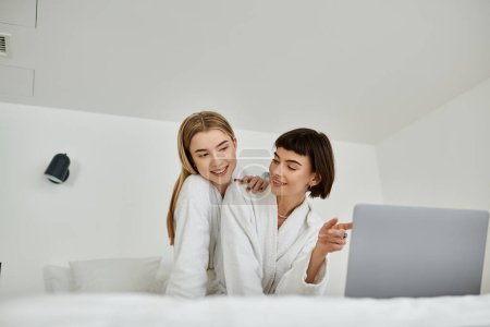 Téléchargez les photos : Un couple lesbien en peignoir se détend sur un lit confortable, partageant un moment tendre de proximité et d'intimité. - en image libre de droit