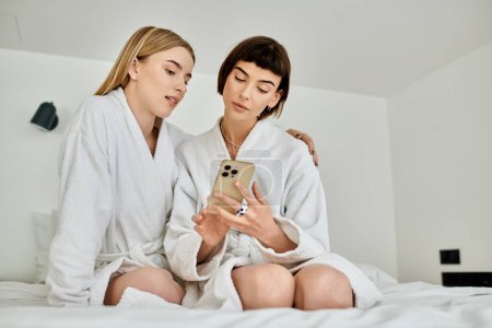 Foto de Dos mujeres con batas de baño sentadas en una cama, absortas en un teléfono celular, compartiendo un momento privado en una habitación de hotel. - Imagen libre de derechos