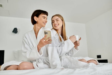 Photo for Beautiful lesbian couple in bath robes relaxing and chatting on top of a bed inside a cozy hotel room. - Royalty Free Image