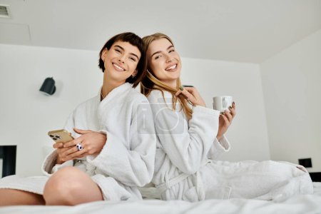 Foto de Una impresionante pareja de lesbianas en batas de baño disfrutar de un momento de relajación mientras se sienta en la parte superior de una cama de lujo dentro de una habitación de hotel. - Imagen libre de derechos