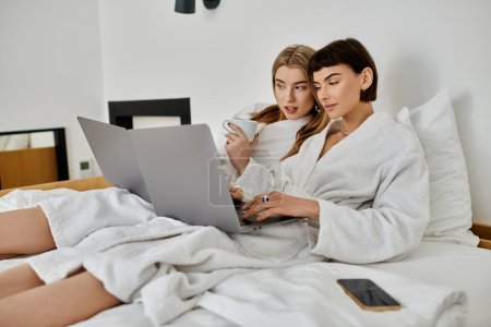 Two women in bath robes sit closely on a bed, focused on a laptop screen in a hotel room.