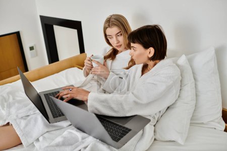 Foto de Dos hermosas mujeres lesbianas en batas de baño sentadas en una cama, cautivadas por la pantalla de un portátil. - Imagen libre de derechos