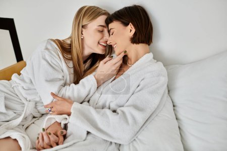 Ein schönes lesbisches Paar in Bademänteln entspannt auf einem Bett, bequem unter einer warmen Decke eingebettet.