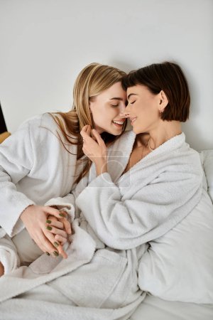 Foto de A serene moment captured as two women, a beautiful lesbian couple, embrace in white robes on a bed inside a hotel room. - Imagen libre de derechos