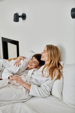 Téléchargez les photos : Deux femmes en peignoirs de bain allongées sur un lit avec draps blancs dans une chambre d'hôtel sereine. - en image libre de droit