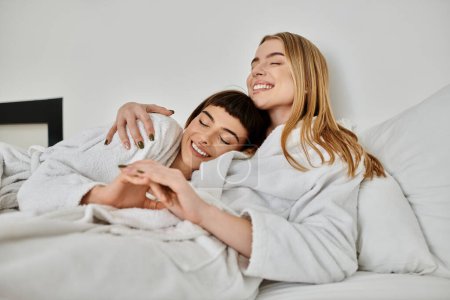Téléchargez les photos : Un beau couple de lesbiennes en peignoirs de bain reposant paisiblement sur un lit confortable à l'intérieur d'une chambre d'hôtel. - en image libre de droit