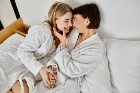 Téléchargez les photos : Un couple lesbien détendu en peignoirs de bain s'assoit sur un lit d'hôtel soigneusement fait, profitant d'un moment de calme ensemble. - en image libre de droit