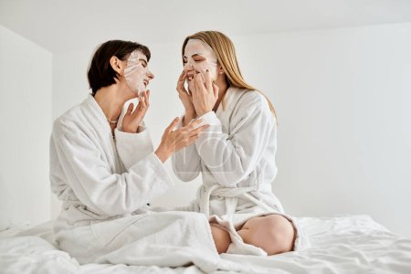 Two women in bath robes sitting on a bed, calmly wearing face masks for a spa-like moment of relaxation.