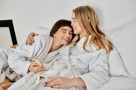 A lesbian couple in bath robes relax, laying in bed under a cozy blanket in a hotel room.