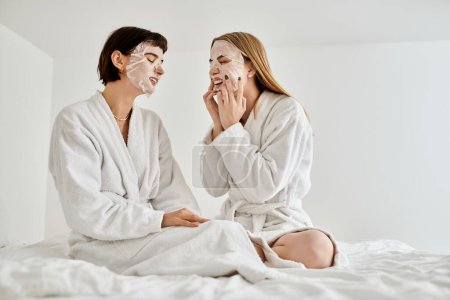 Photo for A beautiful lesbian couple in sheet masks and bath robes peacefully sitting on a luxurious hotel bed together. - Royalty Free Image