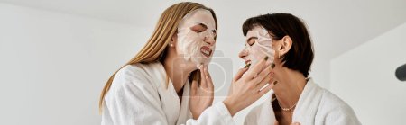 Foto de Dos hermosas mujeres en batas de baño relajándose con máscaras faciales en sus caras en una acogedora habitación de hotel. - Imagen libre de derechos