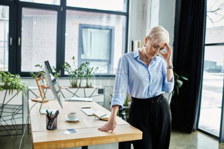 A middle-aged businesswoman in a blue shirt stands at a desk in an office, feeling unwell during menopause.