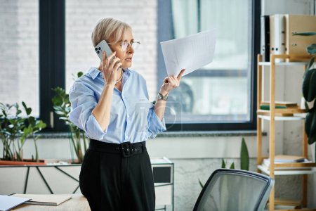 Una mujer de negocios de mediana edad con pelo corto multitarea, hablando por teléfono mientras sostiene un pedazo de papel en un entorno de oficina.