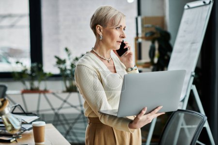 Foto de Una mujer de negocios de mediana edad con el pelo corto habla en un teléfono celular mientras sostiene un ordenador portátil en una oficina moderna. - Imagen libre de derechos