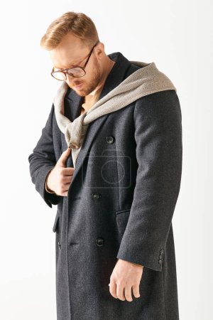A handsome man in a coat and sweater striking a pose against a white wall.