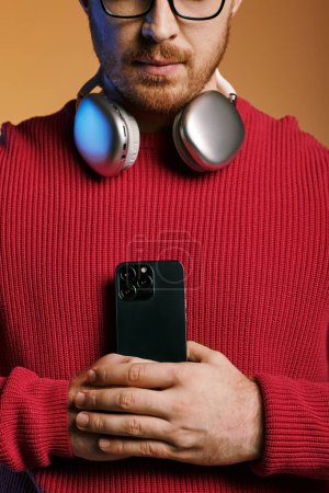 A man in stylish attire listens to music through headphones while holding a cell phone.