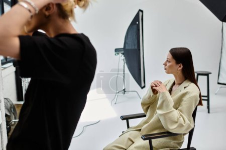 A woman sits near her makeup artist in photo studio.
