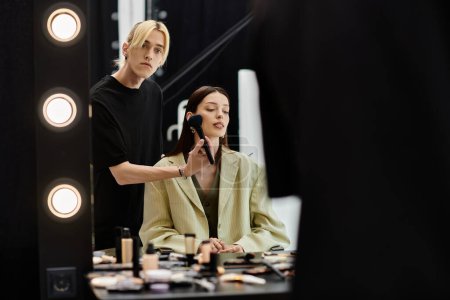 A woman sits before a mirror, her makeup being expertly applied by a makeup artist.