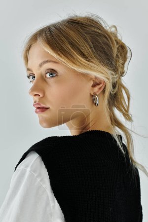 A stylish young woman with blonde hair wearing a black and white sweater and earrings against a grey backdrop.