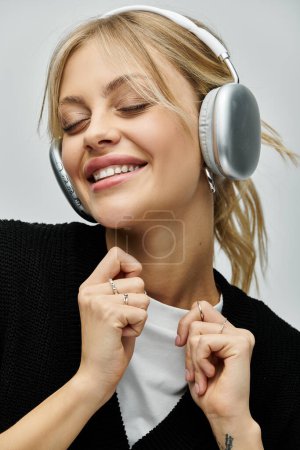 A young woman with blonde hair smiling, wearing stylish attire and headphones, immersed in music on a grey background.