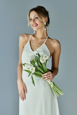Elegant blonde woman in white dress holding a vibrant bouquet of flowers on a grey background.