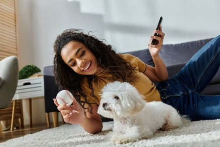 A woman lounging with her bichon frise and checking her phone.