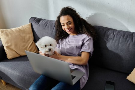Photo for A young woman sitting on a couch, typing on a laptop, accompanied by her fluffy Bichon Frise dog. - Royalty Free Image