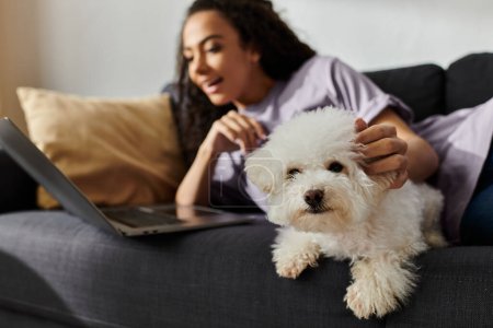 A woman sitting on a couch with her bichon frise dog and a laptop.