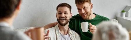 A gay couple smiles and holds mugs while sitting on a couch in their home, likely meeting with family.
