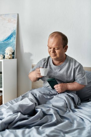 Ein Mann mit Inklusivität sitzt im Bett, hält eine Tasse Kaffee und sein Telefon in der Hand und genießt einen entspannten Morgen.
