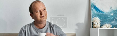 Photo for A man with inclusivity sits in a room, holding a cup, and looks up, lost in thought, with a painting of crashing waves in the background. - Royalty Free Image