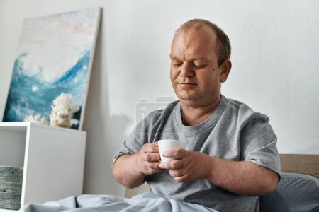 A man with inclusivity sits in bed, holding a cup of coffee in his hands.