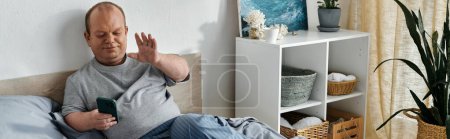 Un homme inclusif s'assoit sur son lit, souriant et saluant tout en tenant un smartphone.
