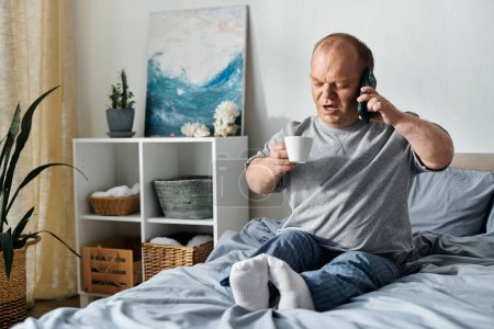 Un hombre con inclusividad se sienta en su cama, sosteniendo una taza de café y hablando por teléfono.