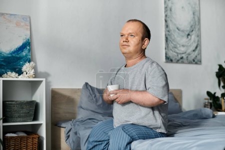 Un homme avec inclusivité est assis sur son lit le matin, tenant une tasse et regardant pensivement hors du cadre.