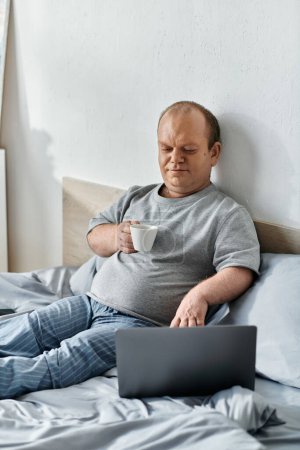 Ein Mann mit Inklusivität sitzt im Bett und genießt eine Tasse Kaffee, während er an seinem Laptop arbeitet.