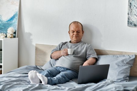 Ein Mann mit Inklusivität sitzt im Bett und genießt eine Tasse Kaffee, während er seinen Laptop benutzt.