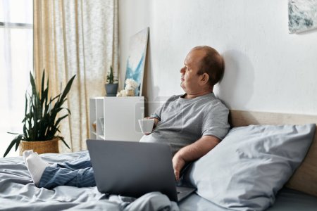 Un homme inclusif est assis dans son lit, tenant une tasse et regardant par la fenêtre, avec un ordinateur portable reposant sur ses genoux.