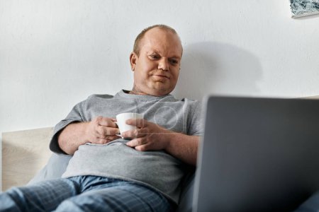Ein inklusiver Mann sitzt auf einem Bett, hält eine Kaffeetasse in der Hand und blickt auf einen Laptop..