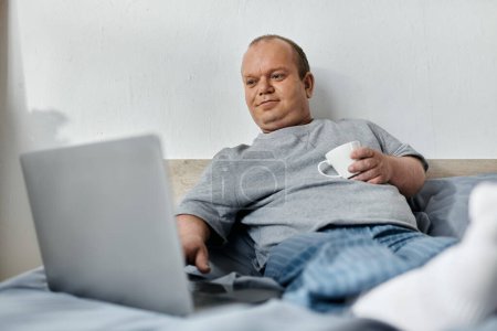 Un hombre con inclusividad se sienta en una cama, relajado y mirando un portátil. Él sostiene una taza blanca en una mano y tiene los pies en alto.