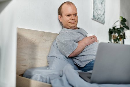 Un homme avec inclusivité s'assoit dans son lit, prend une tasse de café et travaille sur son ordinateur portable.